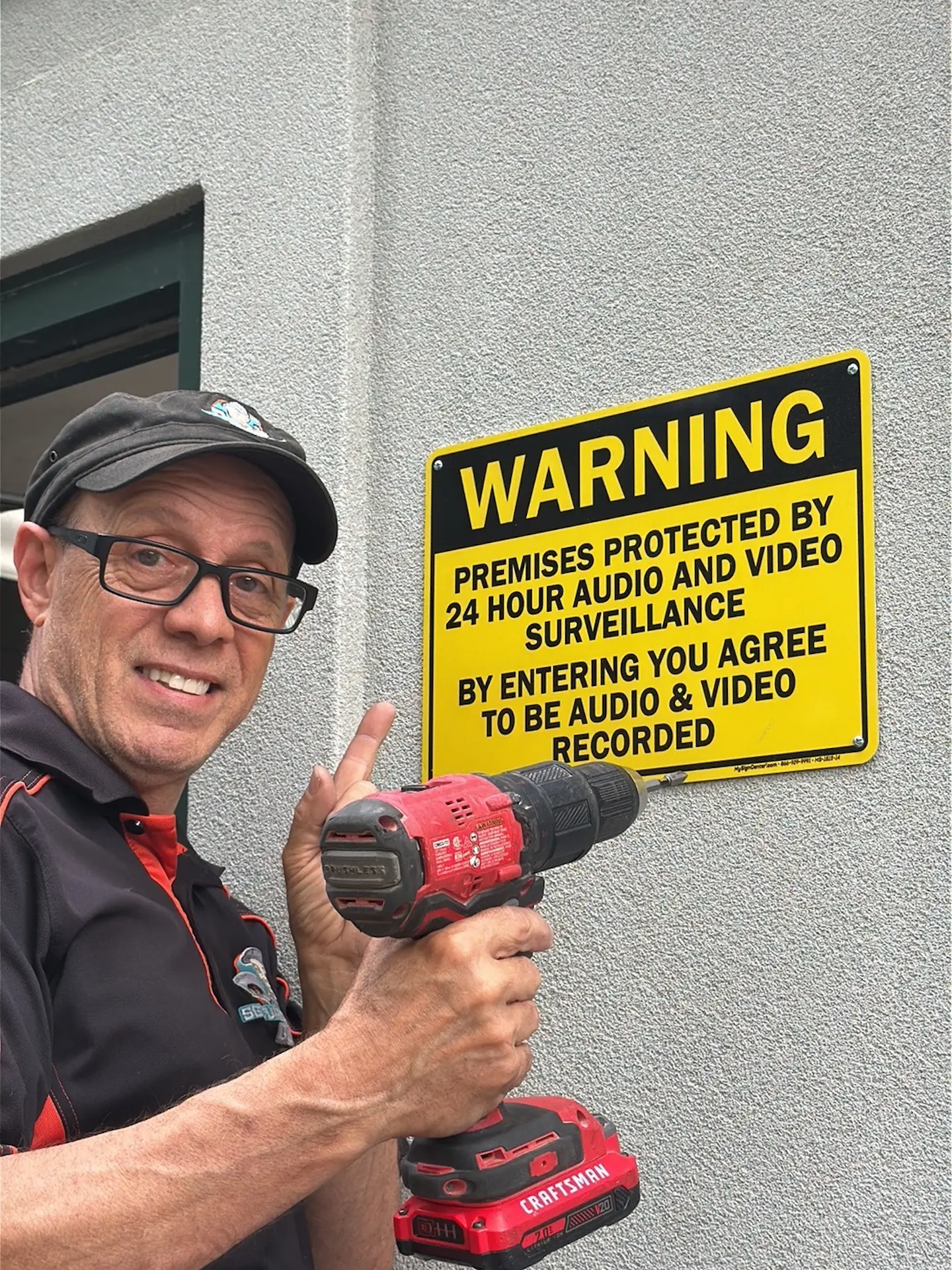 Roland Rowlings of Security Camera Guru mounting a yellow Premises Protected by 24 Hour Audio and Video Surveillance warning sign on the exterior of a Metro Detroit commercial building — the deterrence step that comes with every commercial camera install